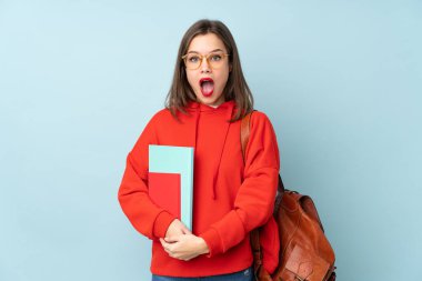 Mavi arka planda, sürpriz bir yüz ifadesiyle izole edilmiş kitapları tutan öğrenci kız.