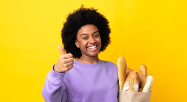 Genç Afrikalı Amerikalı bir kadın sarı arka planda izole edilmiş bir şey alıyor. Başparmakları havada çünkü iyi bir şey oldu.