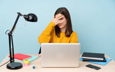 Öğrenci kız evinde çalışıyor. Mavi arka planda izole edilmiş. Gözleri elleriyle kaplıyor ve gülümsüyor.