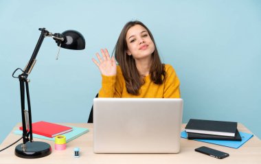 Evinde okuyan öğrenci kız mavi arka planda mutlu bir ifadeyle selamlıyor.