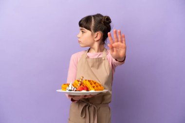 Mor arka planda izole edilmiş waffle tutan küçük beyaz kız dur hareketi yapıyor ve hayal kırıklığına uğruyor.