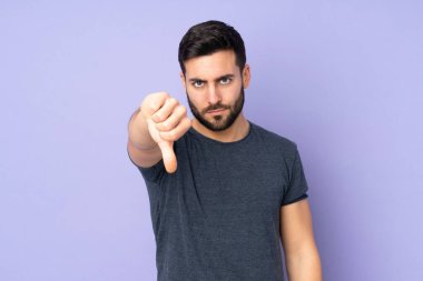 Kafkasyalı yakışıklı adam, izole edilmiş mor arka planda negatif ifade ile baş parmağını aşağı gösteriyor.