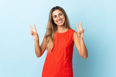 İzole edilmiş mavi arka planda genç İspanyol kadın iki eliyle zafer işareti gösteriyor.
