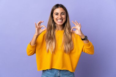 İzole edilmiş mor arka planda genç İspanyol kadın iki eliyle tamam işareti gösteriyor.