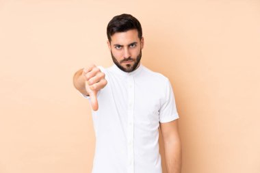 Beyaz tenli yakışıklı adam bej arka planda izole edilmiş, başparmağını negatif ifade ile aşağı gösteriyor.