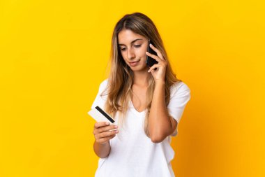 Sarı arka plan izole edilmiş genç İspanyol kadın kredi kartıyla cep telefonundan alışveriş yapıyor.