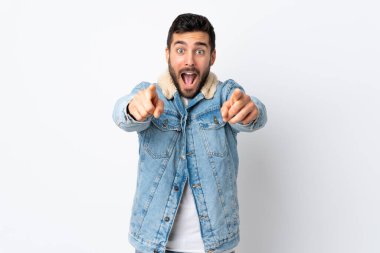 Beyaz arka planda sakallı yakışıklı genç adam şaşırmış ve ön tarafı işaret ediyor.