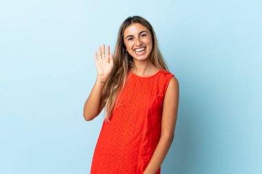 İzole edilmiş mavi arka planda genç İspanyol kadın mutlu bir ifadeyle selamlıyor.