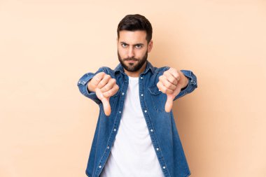 Beyaz tenli yakışıklı adam bej arka planda izole edilmiş, baş parmağını iki eliyle aşağı gösteriyor.