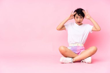Genç Vietnamlı kadın, kısa saçlı, pembe arka planda, yerde oturuyor, sürpriz bir ifadeyle.