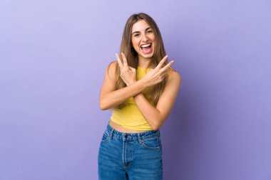 İzole edilmiş mor arka planda gülümseyen ve zafer işareti gösteren genç bir kadın.