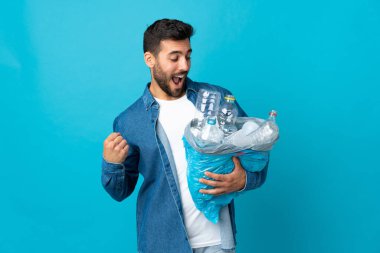 Mavi arka planda izole edilmiş bir şekilde zaferi kutlamak için elinde plastik şişelerle dolu bir çanta tutan genç beyaz bir adam.