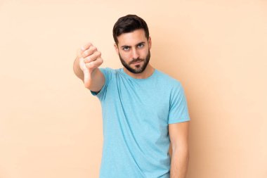 Beyaz tenli yakışıklı adam bej arka planda izole edilmiş, başparmağını negatif ifade ile aşağı gösteriyor.