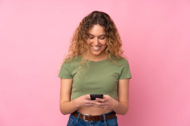 Pembe arka planda izole edilmiş kıvırcık saçlı genç sarışın kadın cep telefonuyla bir mesaj gönderiyor.