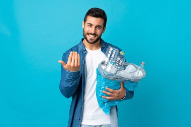 Mavi arka planda izole edilmiş plastik şişelerle dolu bir çanta taşıyan genç beyaz bir adam, bizimle gelmesi için davet ediyor. Gelmene sevindim.