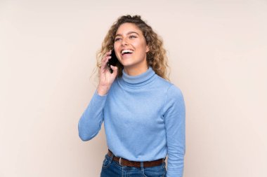 Sarışın, kıvırcık saçlı, balıkçı yaka kazak giyen, bej arka planda izole edilmiş ve cep telefonuyla sohbet eden genç bir kadın.