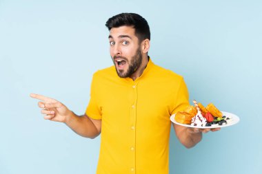 Mavi arka planda izole edilmiş waffle tutan beyaz adam şaşırdı ve parmağını yana çevirdi.