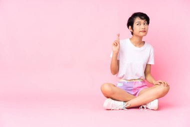 Kısa saçlı genç Vietnamlı kadın pembe arka planda izole bir şekilde yerde oturuyor ve dua ediyor.