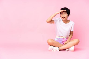 Kısa saçlı genç Vietnamlı kadın, izole edilmiş pembe arka planda yerde oturuyor. Bir şeye bakmak için çok uzaklara bakıyor.