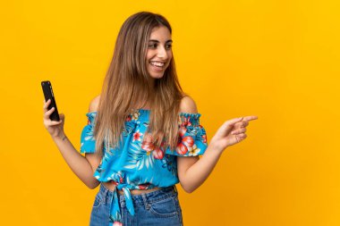 İzole edilmiş sarı arka planda cep telefonu kullanan genç bir kadın bir ürün sunmak için kenarı işaret ediyor.