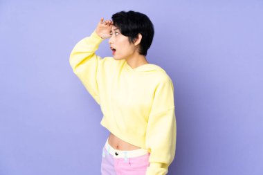 Genç Vietnamlı bir kadın, kısa saçlı, mor arka planda, şaşırmış bir ifadeyle yan tarafa bakıyor.