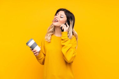 Sarı arka planda izole edilmiş genç bir kız. Elinde kahve ve cep telefonu var.