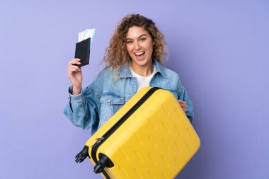 Kıvırcık saçlı genç bir kadın, mor arka planda izole edilmiş, bavul ve pasaportla tatilde.