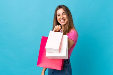 Soyutlanmış mavi arka planı olan genç bir kadın alışveriş torbaları tutuyor ve gülümsüyor.