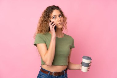 Pembe arka planda izole edilmiş kıvırcık saçlı genç sarışın bir kadın. Elinde kahve ve cep telefonu var.