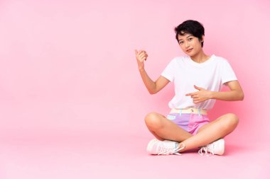 Genç Vietnamlı kadın, kısa saçlı, pembe arka planda, yerde oturmuş gitar hareketi yapıyor.