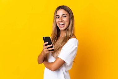 İzole edilmiş sarı arka planı olan genç İspanyol bir kadın cep telefonu tutuyor ve kollarını kavuşturuyor.