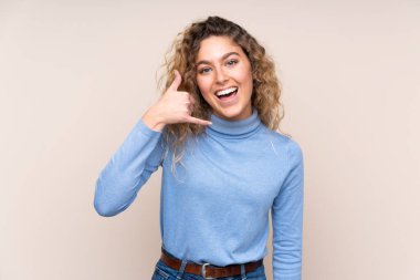 Sarışın, kıvırcık saçlı, balıkçı yaka kazak giyen, bej arka planda izole edilmiş, telefon hareketi yapan genç bir kadın. Bana geri dönüş işareti ver.