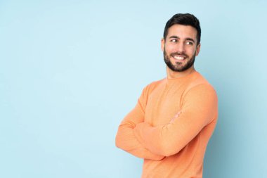 Kollarını kavuşturmuş, mavi arka planda mutlu, beyaz, yakışıklı bir adam.