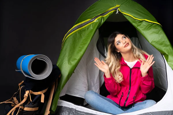 Teenager girl inside a camping green tent isolated on black background making doubts gesture