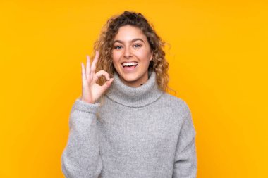 Sarışın, kıvırcık saçlı, balıkçı yaka kazak giyen, sarı arka planda izole edilmiş, şaşırmış ve iyi bir işaret gösteren genç bir kadın.