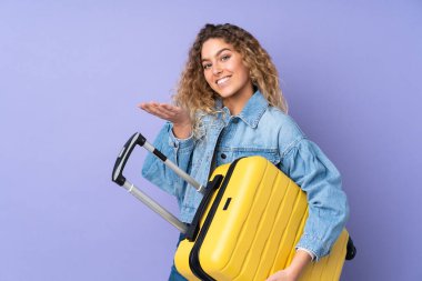 Kıvırcık saçlı genç bir kadın, mor arka planda izole edilmiş seyahat valiziyle tatilde.