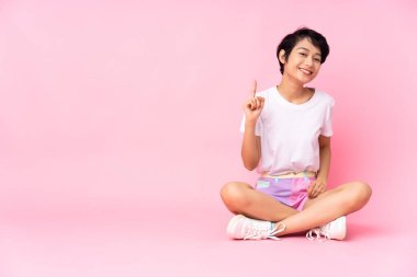 Kısa saçlı genç Vietnamlı kadın pembe arka planda izole edilmiş bir şekilde yerde oturuyor ve en iyinin işareti olarak parmağını kaldırıyor.