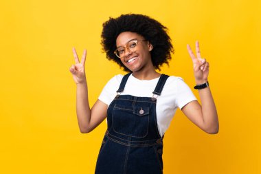 Sarı arka planda izole edilmiş genç Afrikalı Amerikalı kadın iki eliyle zafer işareti gösteriyor.