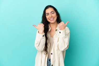 Mavi arka planda izole edilmiş genç beyaz kadın başparmakları havada ve gülümsüyor.