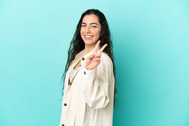 Mavi arka planda izole edilmiş genç beyaz kadın gülümsüyor ve zafer işareti gösteriyor.