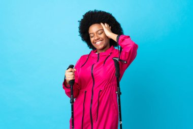 Sırt çantalı ve yürüyüş direkleriyle mavi arka planda izole edilmiş genç Afrikalı Amerikalı kahkahalar