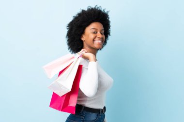 Genç Afrikalı Amerikalı bir kadın mavi arka planda izole edilmiş alışveriş torbaları tutuyor ve gülümsüyor.