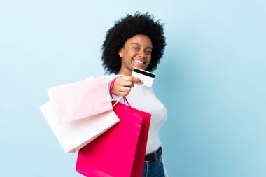 Genç Afrikalı Amerikalı bir kadın mavi arka planda izole edilmiş. Elinde alışveriş çantaları ve bir kredi kartı var.