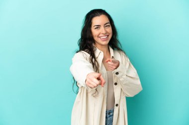 Mavi arka planda izole edilmiş genç beyaz kadın şaşırmış ve ön tarafı işaret ediyor.