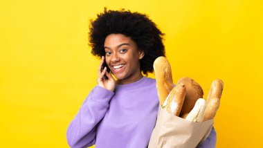Genç Afrikalı Amerikalı bir kadın sarı arka planda izole edilmiş bir şey alıyor cep telefonuyla sohbet ediyor.
