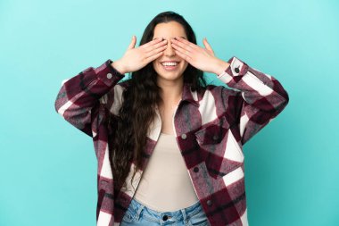 Mavi arka planda izole edilmiş genç beyaz bir kadın. Gözleri elleriyle kaplıyor ve gülümsüyor.