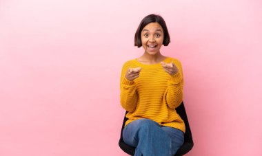 Genç melez kadın pembe arka planda izole bir sandalyede oturuyor şaşırmış ve ön tarafı işaret ediyor.