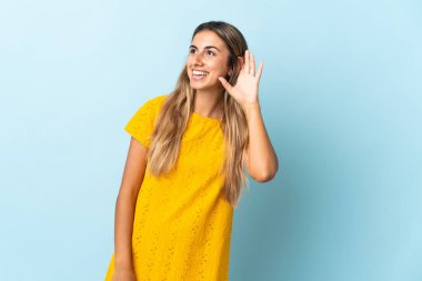 İzole edilmiş mavi arka planda genç bir İspanyol kadın. Kulağına elini sürerek bir şeyler dinliyor.