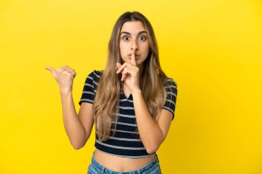 Sarı arka planda izole edilmiş genç, beyaz bir kadın. Yan tarafı işaret ediyor ve sessizlik hareketi yapıyor.