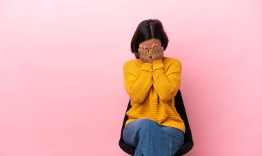 Genç melez kadın pembe arka planda izole edilmiş bir sandalyede oturuyor gözlerini elleriyle kapatıyor.